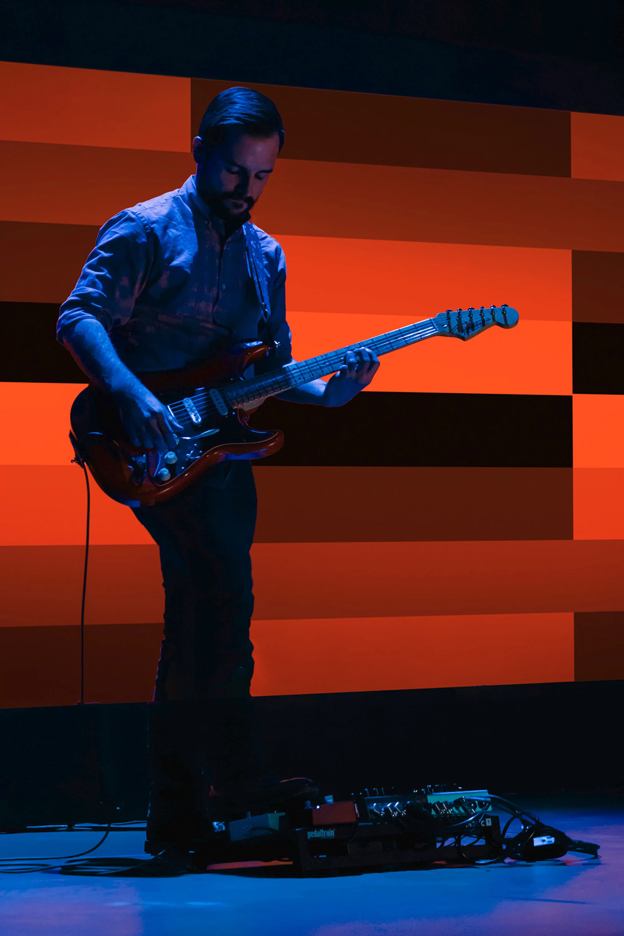A man with a beard stands playing an electric guitar on a stage with orange, white, and black geometric panel backdrop, bathed in blue light.