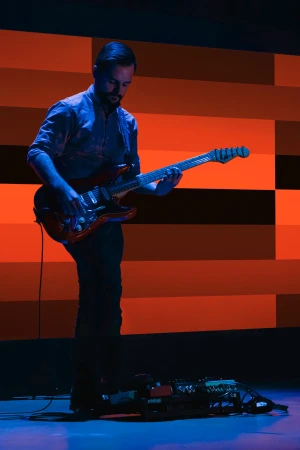 A man with a beard stands playing an electric guitar on a stage with orange, white, and black geometric panel backdrop, bathed in blue light.