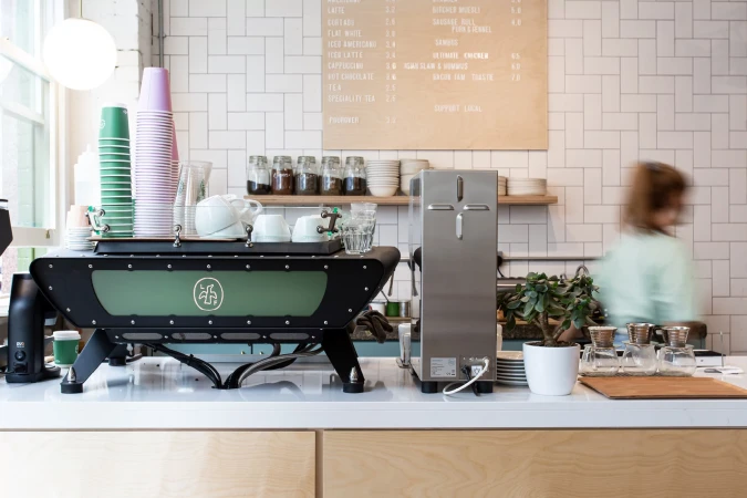Coffee machine and cups at cafe. Espresso maker, paper cups, clear glasses, and barista blurred in background inside cafe.