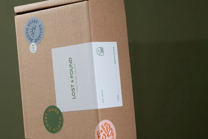Tan corrugated cardboard box with food company label, 'Lost & Found: Fresh Coffee, Fine Eats' and round stickers on a green background.