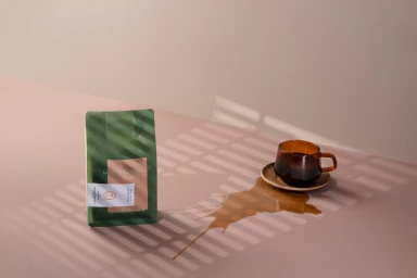 Green coffee bag and a glass cup of coffee with a spill on a warm surface under striped window shadows.