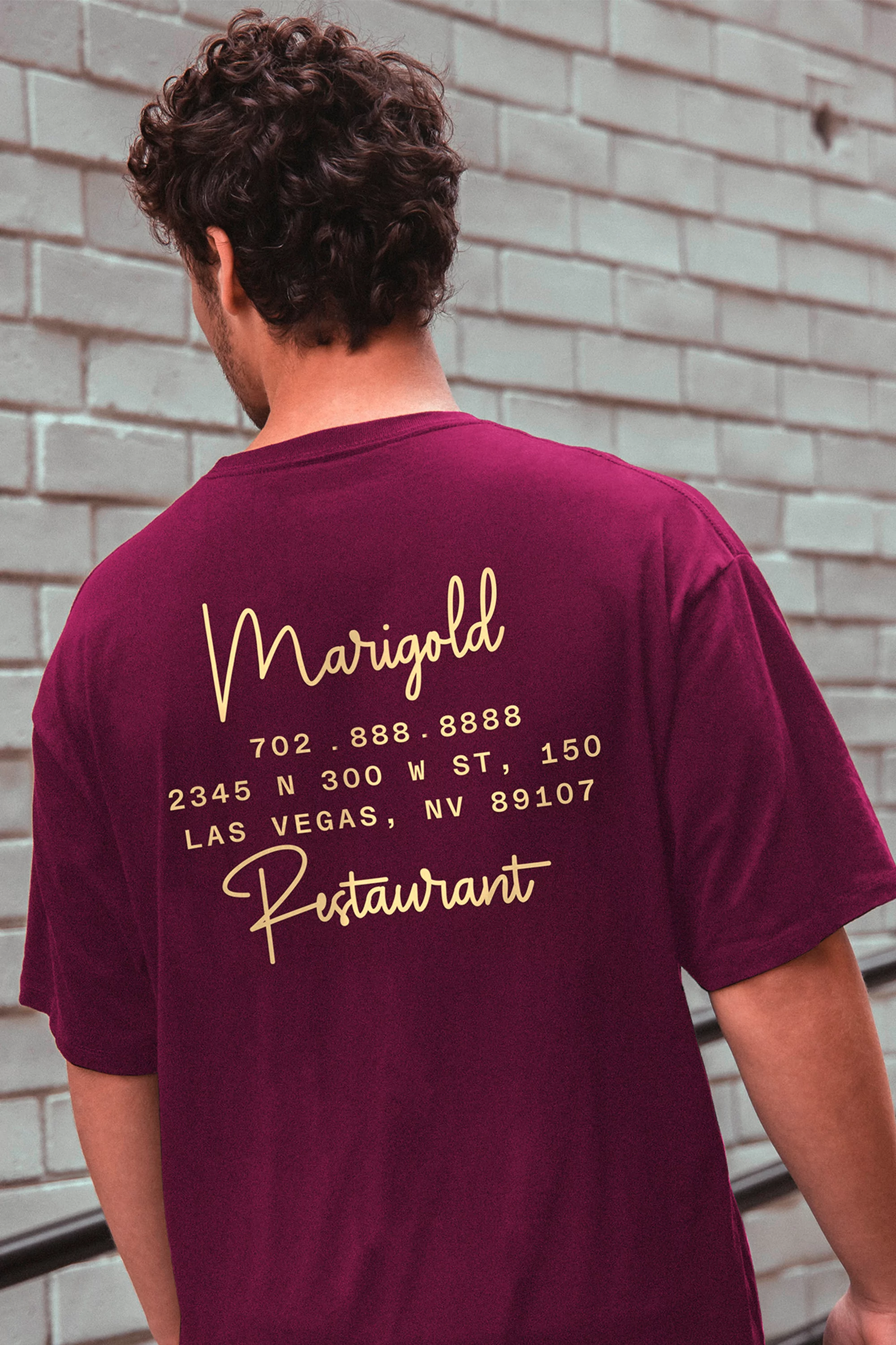 A man in a plum T-shirt stands against a brick wall with Marigold Restaurant contact information printed on the back.