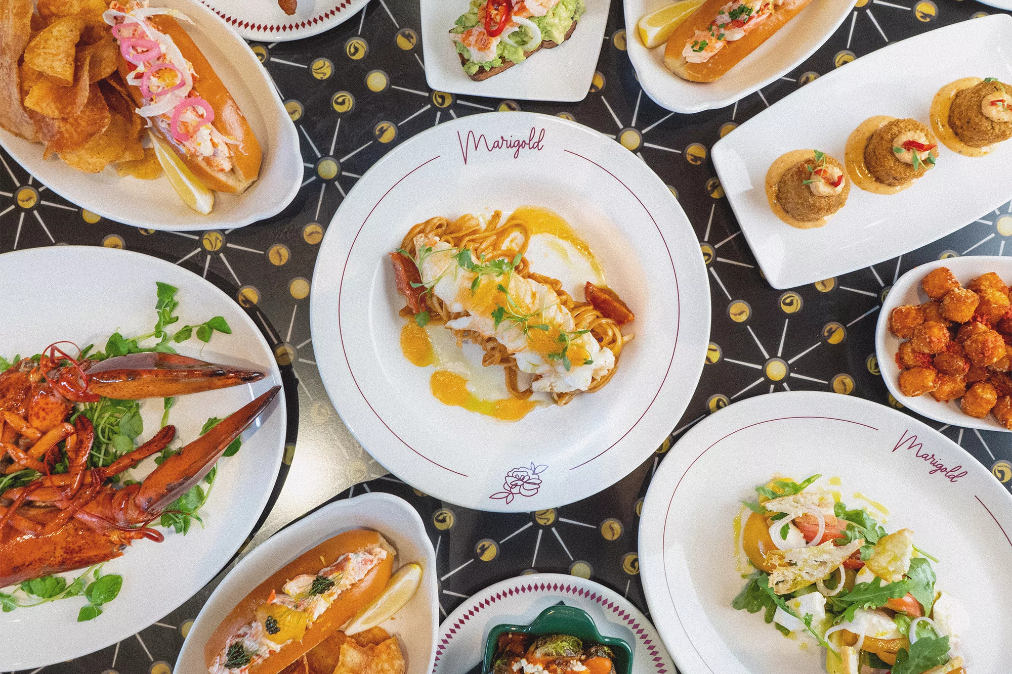 Overhead shot of a restaurant table featuring plates with lobster rolls, pasta, toast, salad, and fried food, all on a patterned table.