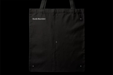 A black tote bag against a solid black background featuring white branding text and alignment crop marks.