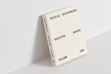 Hardcover monograph book titled Nicolas Schuybroek Selected Works Volume One leaning against a minimal white corner wall.