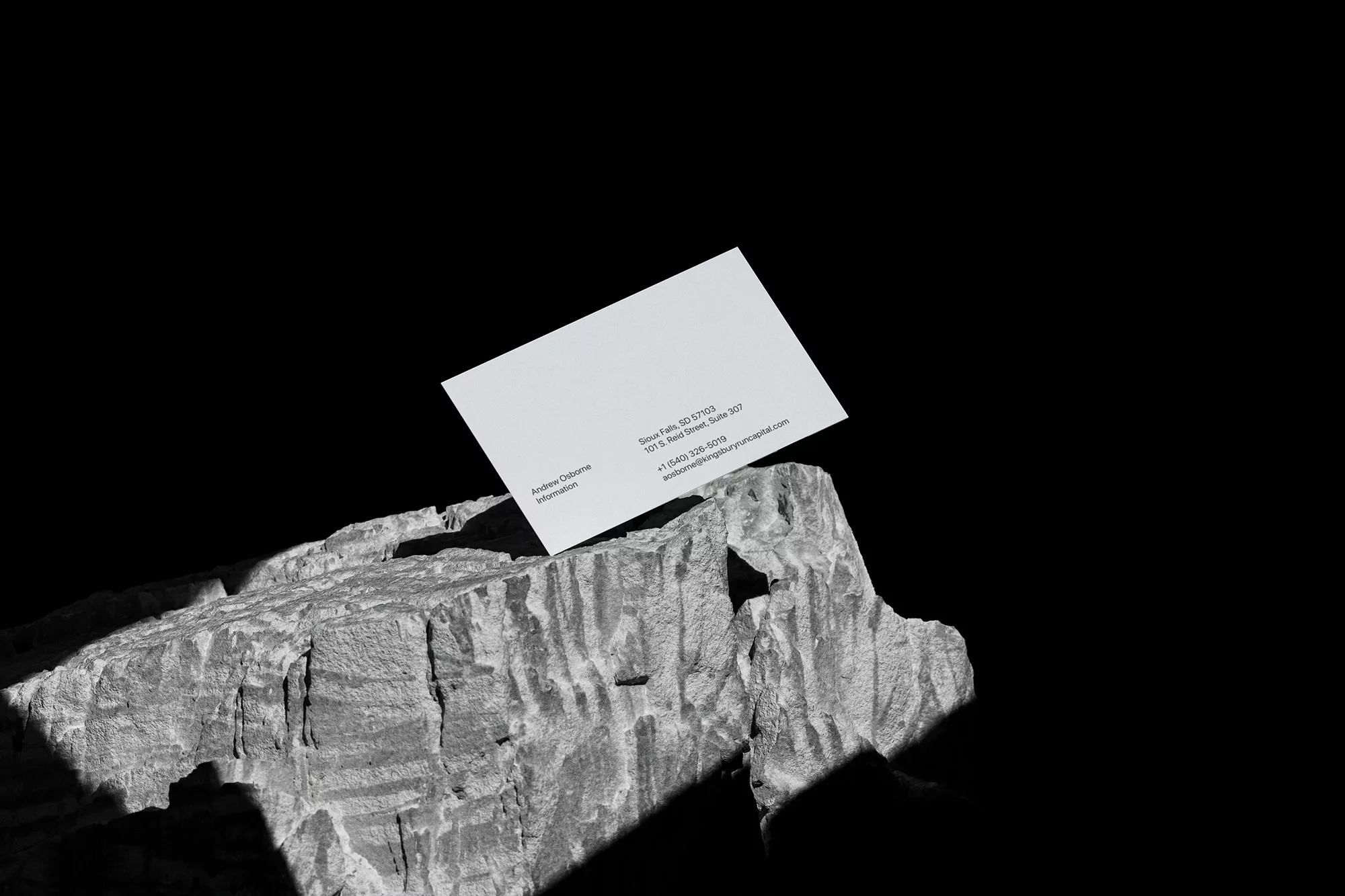 Elevated view of a business card atop a textured rock against a stark black backdrop, featuring company contact details and logo.