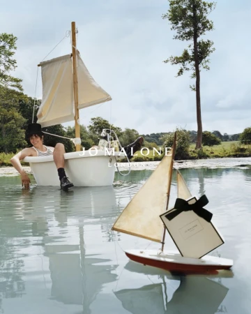 Child reclines in bathtub sailboat on a lake, Jo Malone gift, capturing whimsical outdoor beauty and youthful nautical themes.