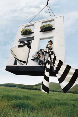 Elegant fashion shot featuring a woman in a black-and-white dress sitting on a building facade suspended over a green field under a cloudy sky.