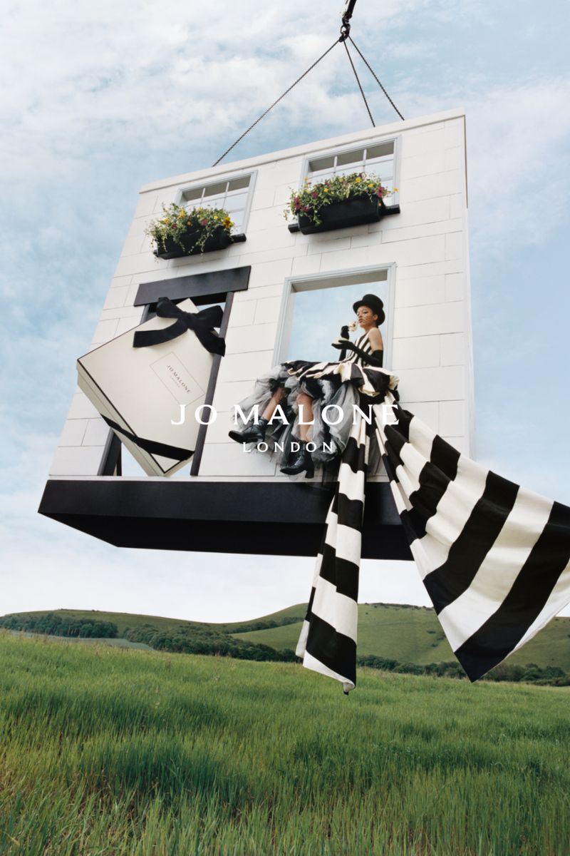 Perfume ad with a fashion model in tulle dress sitting in a window on a suspended building section with a Jo Malone box and green field.