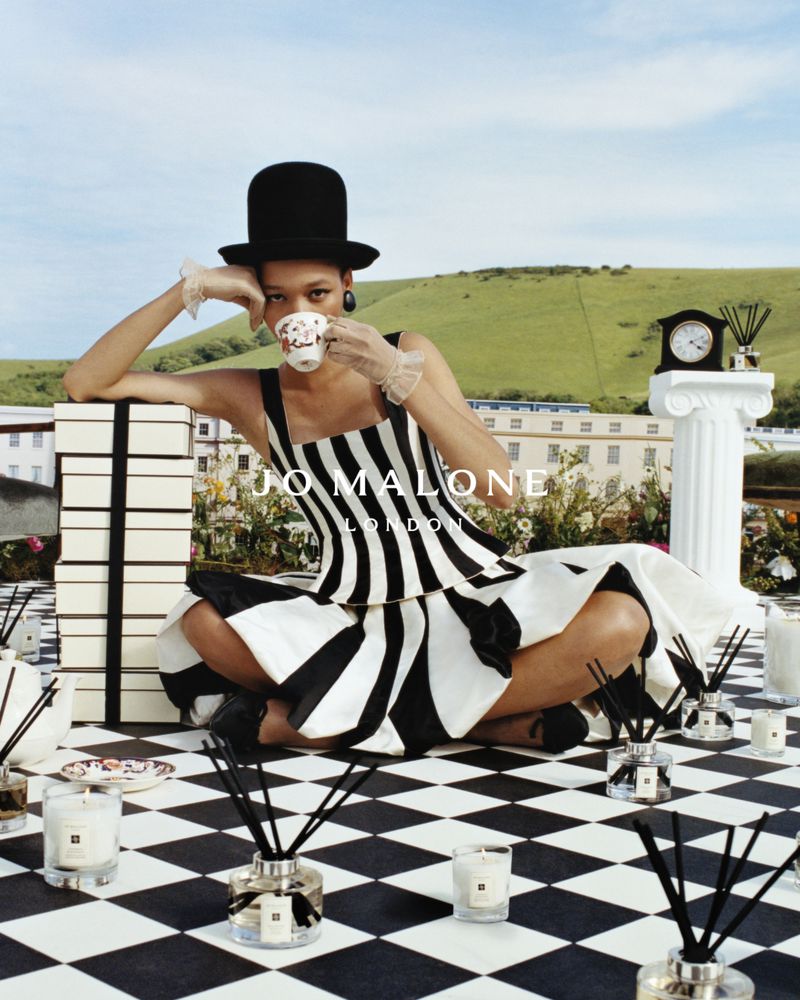 A woman in a black and white striped dress and top hat poses in a whimsical outdoor setting with Jo Malone London perfume.