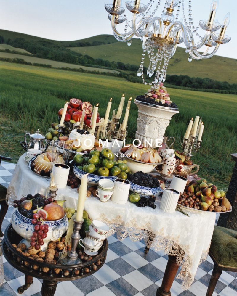 Opulent outdoor feast with fruit platters, candelabras, and fine china on a checkered floor, set against a lush green hill backdrop.