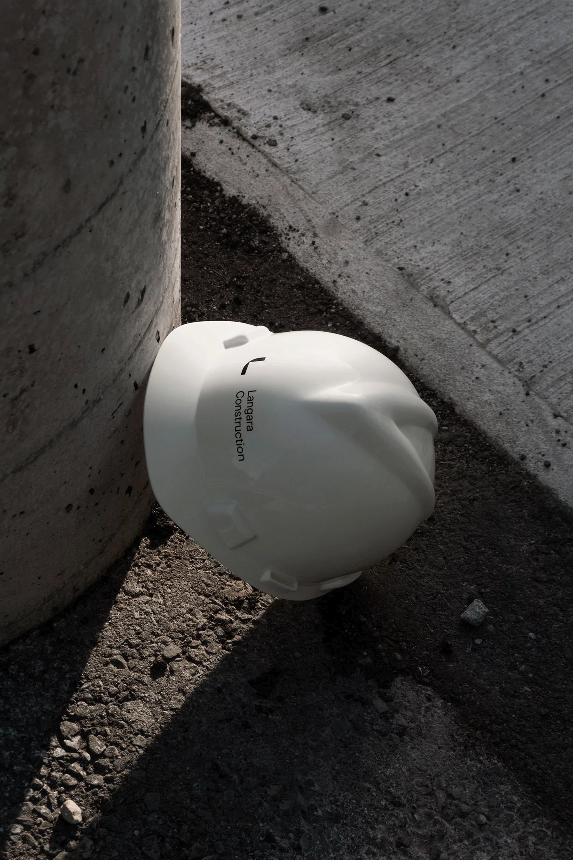 Hard hat rests on the ground next to a concrete pillar in an outdoor setting with shadows and construction industry connotations.