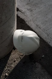 White hard hat resting against a concrete pillar on rough stone ground under natural sunlight.