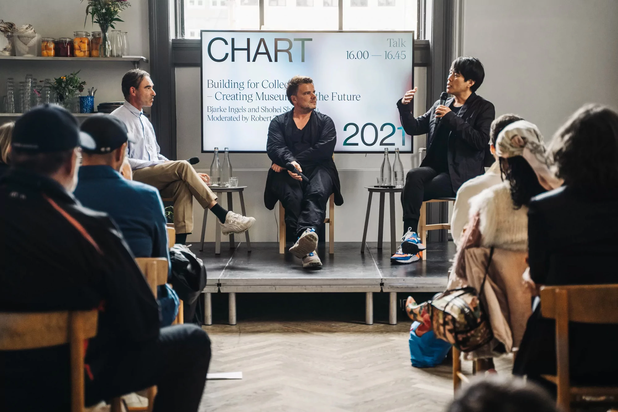 A panel discussion featuring three speakers on a stage, addressing an audience at an event with a screen displaying 'CHART' and event details.
