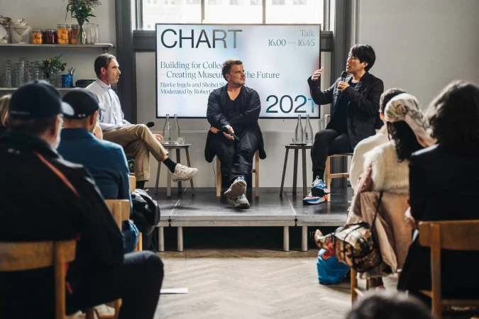 A panel discussion featuring three speakers on a stage, addressing an audience at an event with a screen displaying 'CHART' and event details.