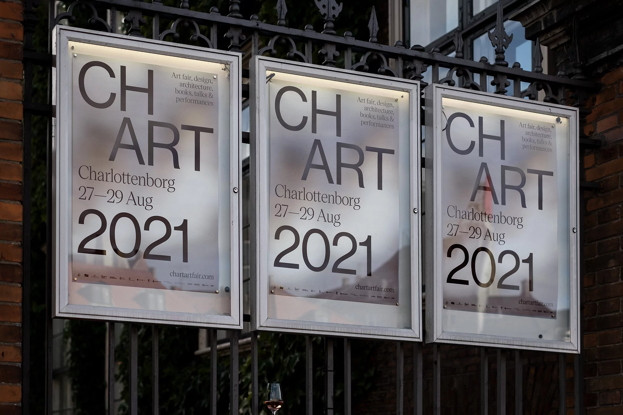 Three illuminated CH ART posters on a dark metal gate announce the Charlottenborg art fair, design, architecture, and performance event in 2021.