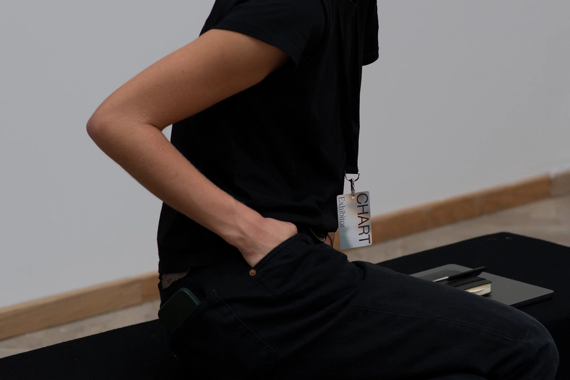 Unidentifiable person with hand in pocket, wearing black shirt and pants, and an exhibitor badge, seated on a black bench.