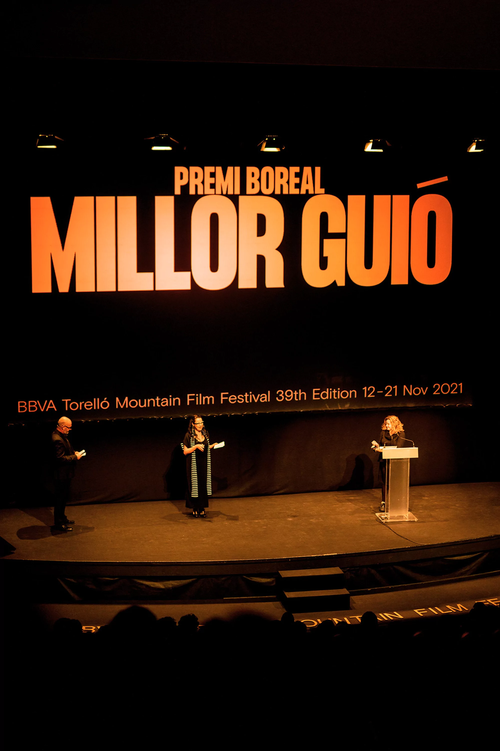 Panel discussion at the Torelló Mountain Film Festival features three speakers on stage under event signage in a darkened auditorium.