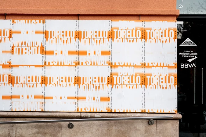Outdoor posters on a building wall promote the Torelló Mountain Film Festival with bold typography and orange accents, near a glass door entrance.