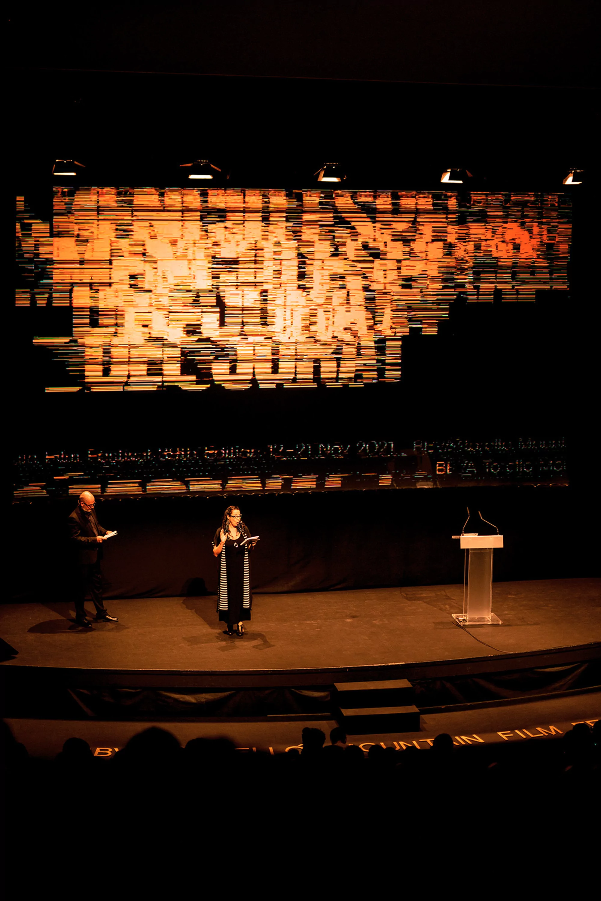 Film festival event: A woman speaks on stage with a man at her side, a podium, and a large screen in the background.