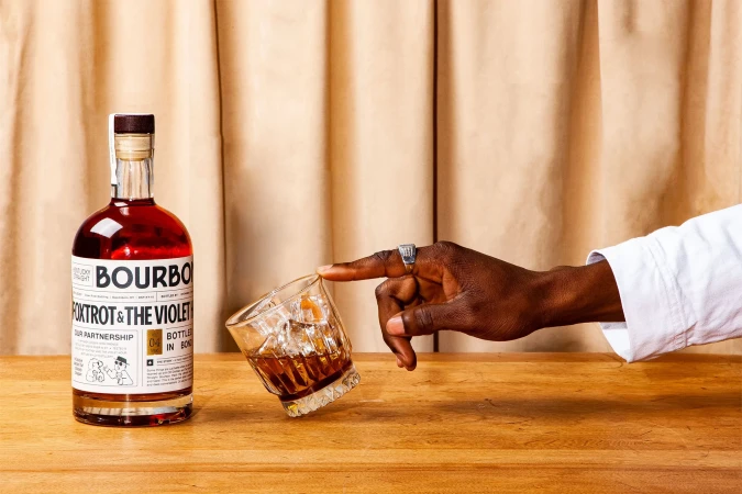 Bourbon bottle and an old-fashioned cocktail on a wooden table with a hand touching the glass, against a neutral draped background.