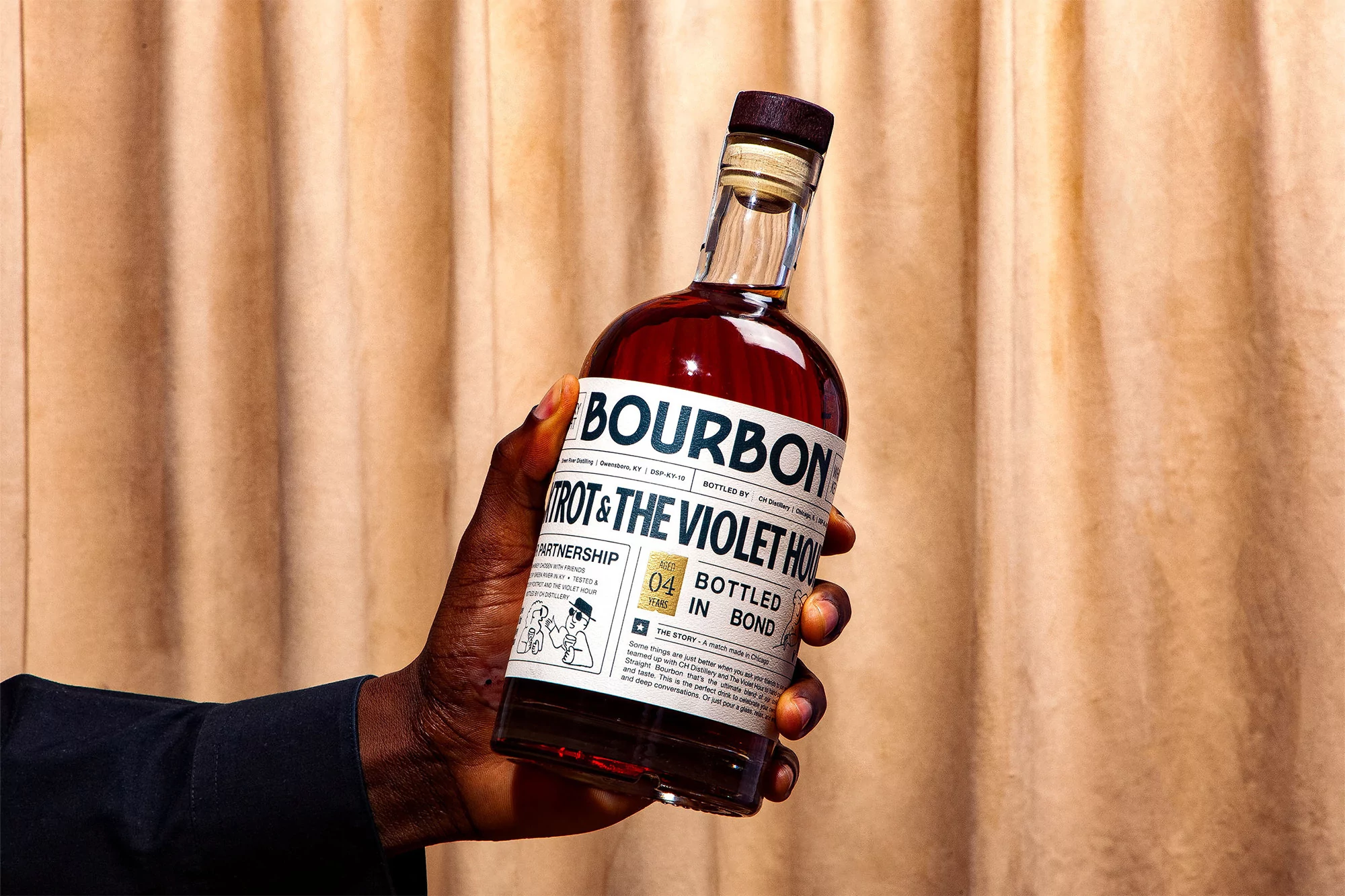 Close-up of a dark-skinned hand holding a bottle of bourbon against a neutral curtain. The bottle has a white label.