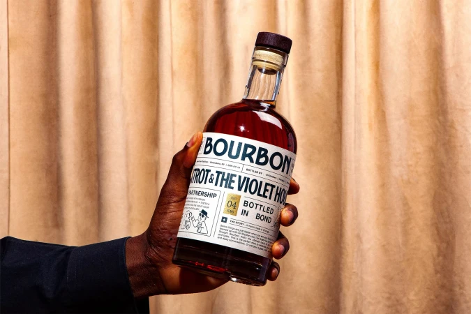 Close-up of a dark-skinned hand holding a bottle of bourbon against a neutral curtain. The bottle has a white label.