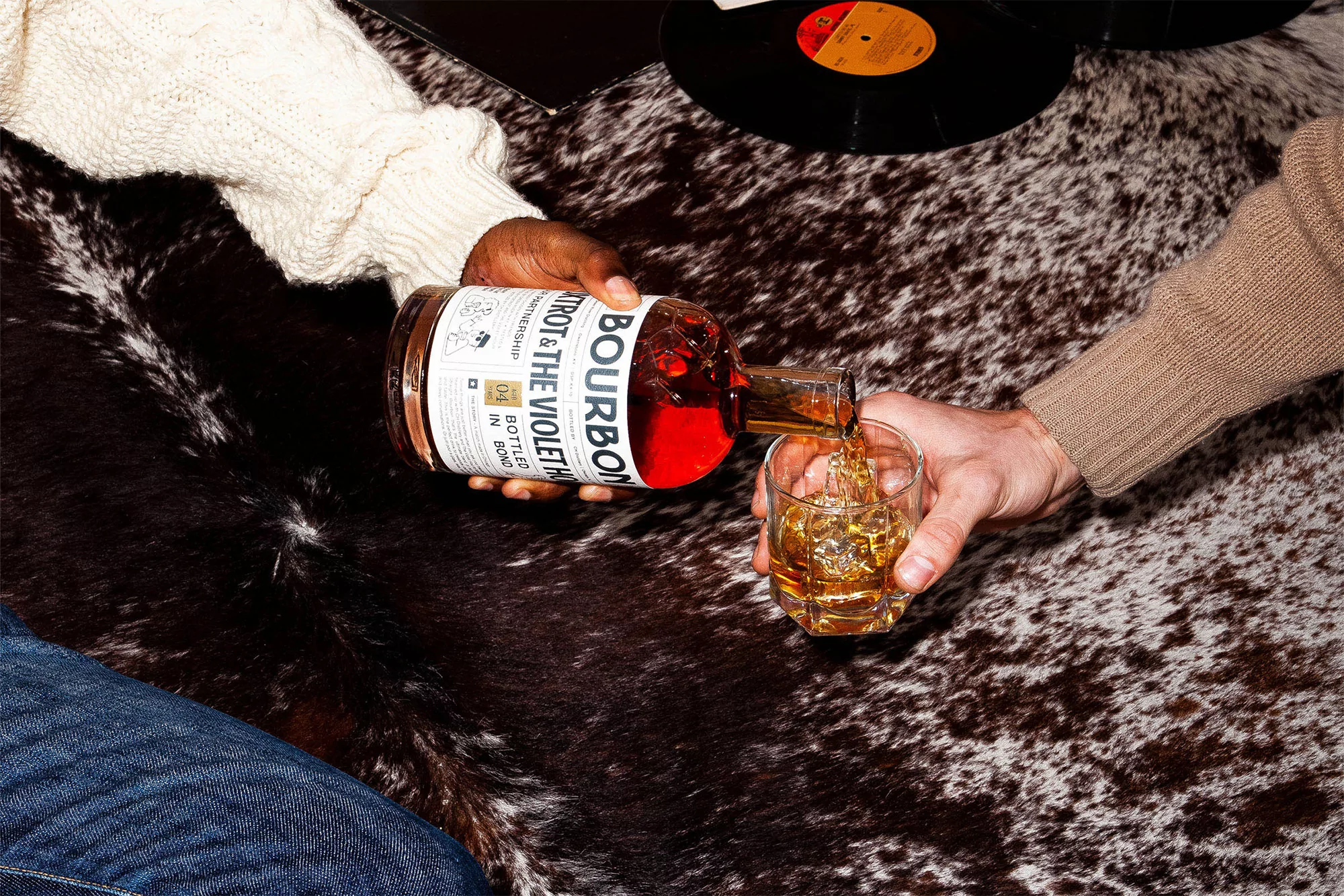 Bourbon being poured into a glass by two people wearing sweaters on a cowhide rug with vinyl records nearby. 