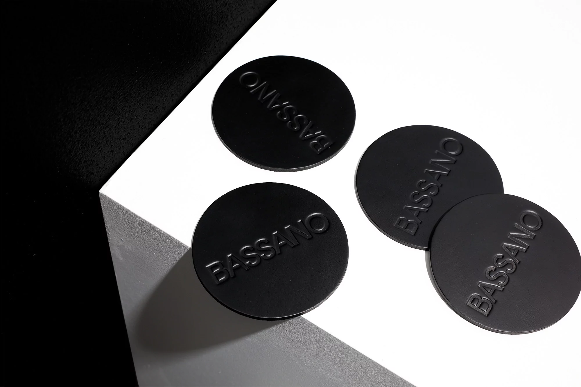 Four black circular product lids with 'BASSANO' embossed sit on a white surface against a dark backdrop in a studio product shot.