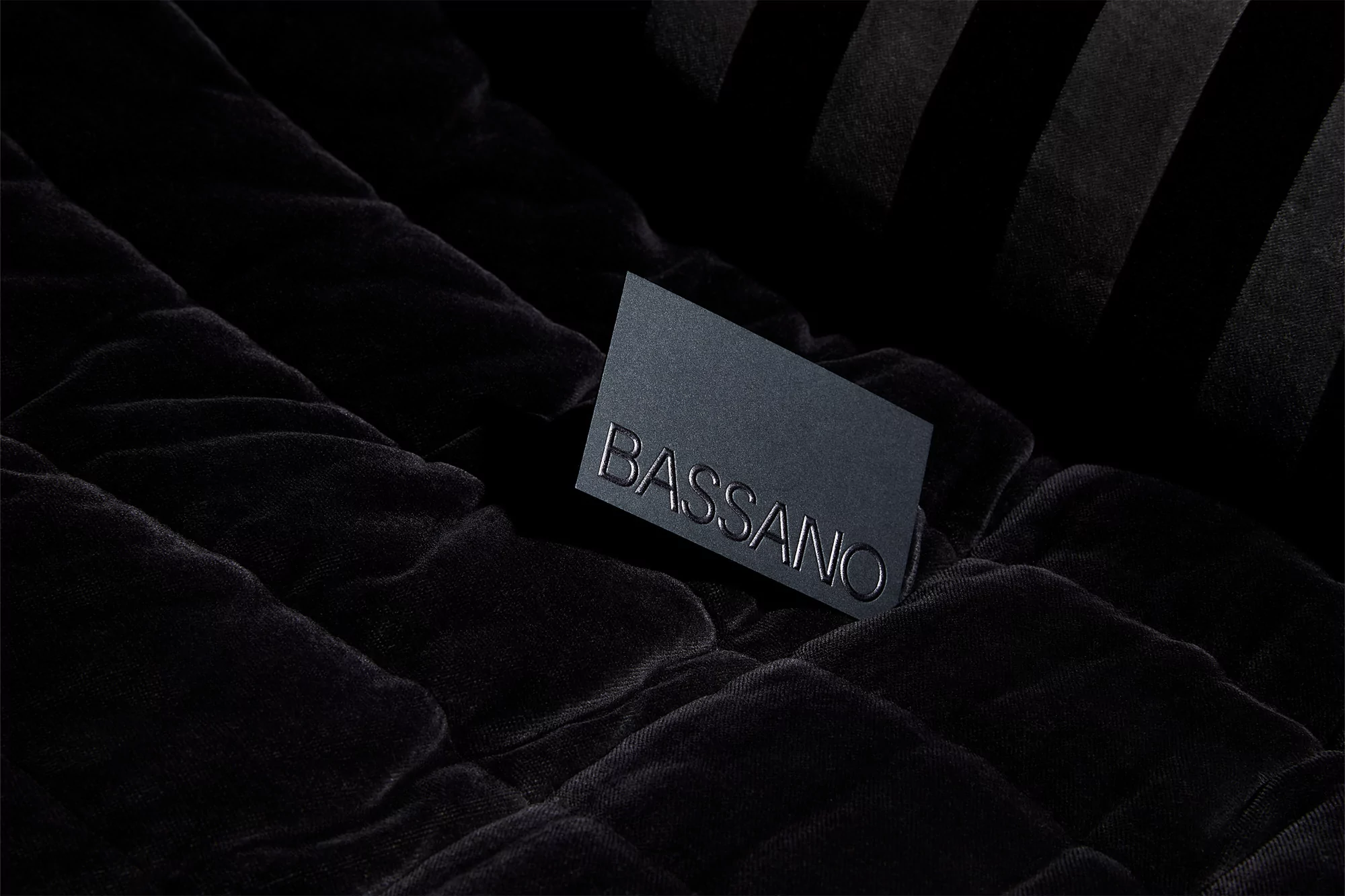 Close-up of a black business card with the text BASSANO on a dark, crumpled fabric backdrop; dramatic low-key lighting.