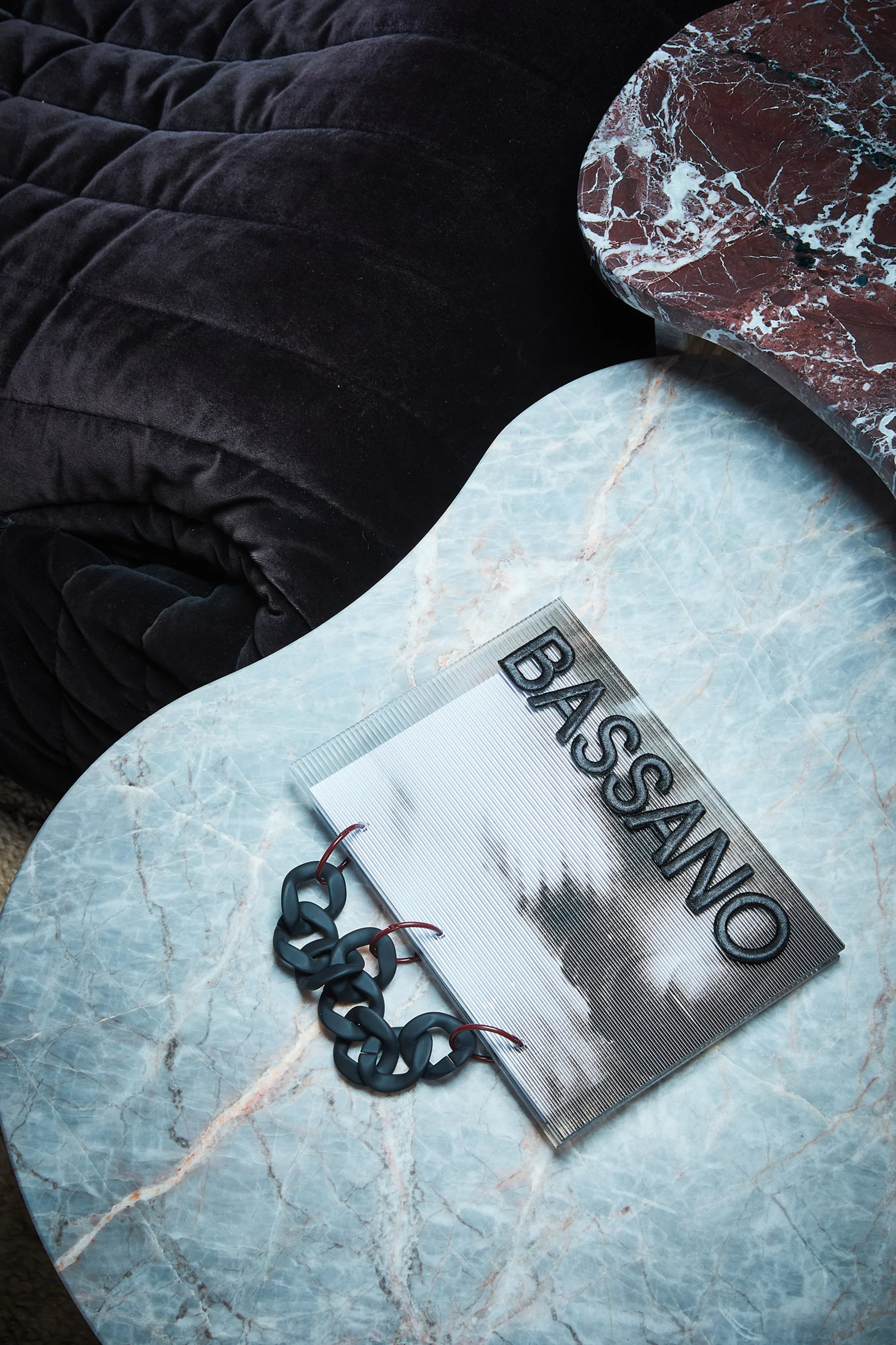 Still-life shot of a Bassano notebook with chain detail on a marble table, alongside dark, plush fabric and reddish marble.