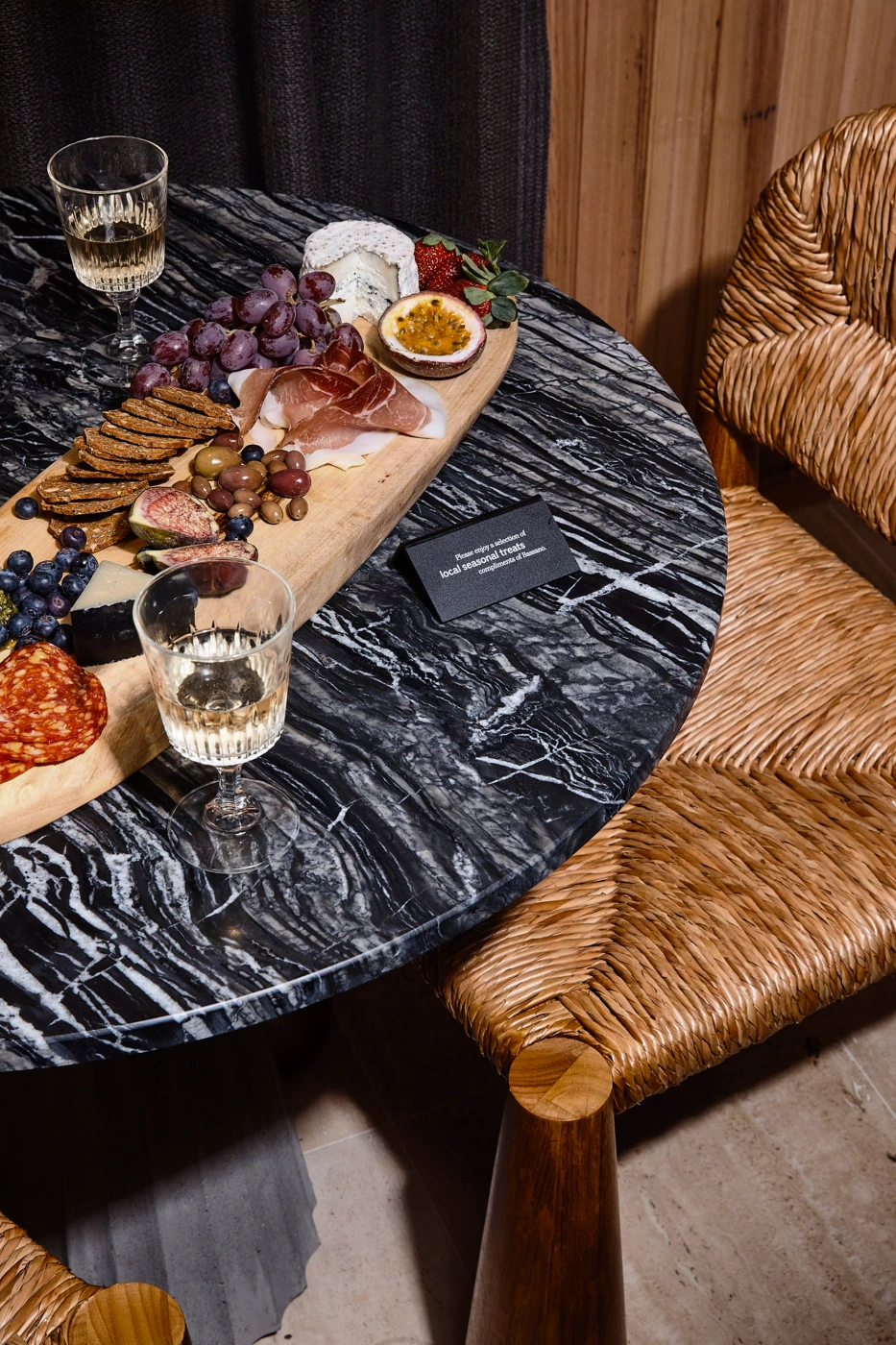 Charcuterie board with cheeses, grapes, and crackers on a black marble table next to woven rattan chairs