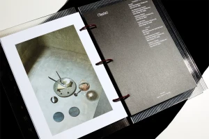Open ring binder with a printed photo showing cheese, wine, and Bassano coasters alongside a grey text page.