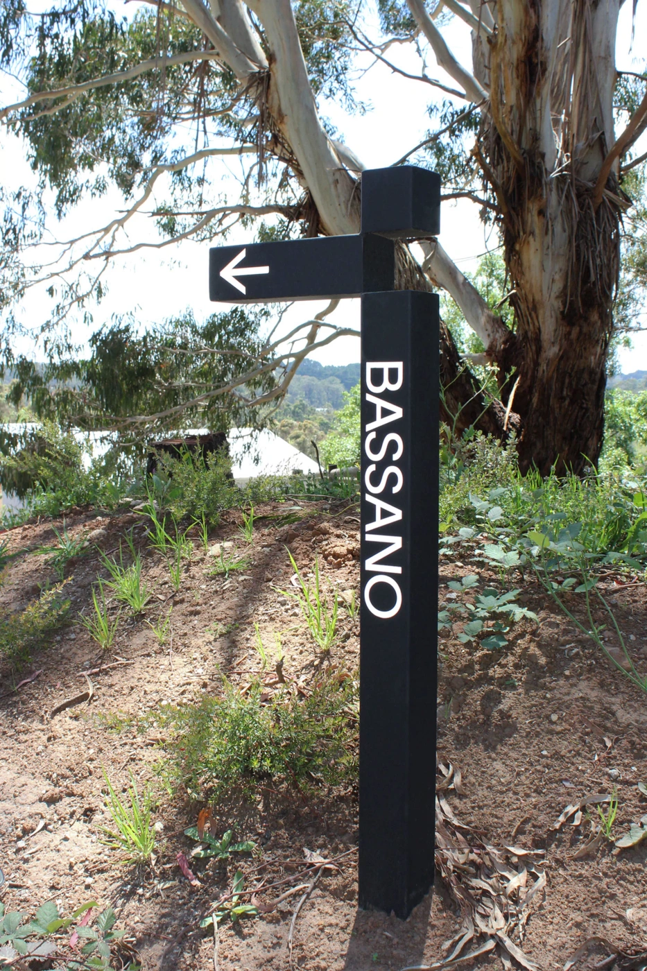 A black directional sign with white text reading BASSANO stands outdoors in a natural landscape with trees.