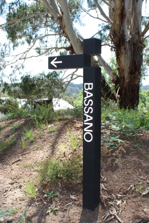 A black directional sign with white text reading BASSANO stands outdoors in a natural landscape with trees.