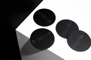 Four round black Bassano coasters arranged on a sharp geometric white and dark surface in a high-contrast minimalist composition
