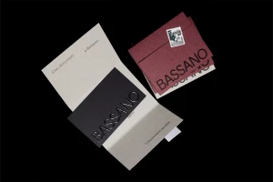 Bassano brand identity stationery featuring folded paper cards and burgundy envelopes on dark surface.