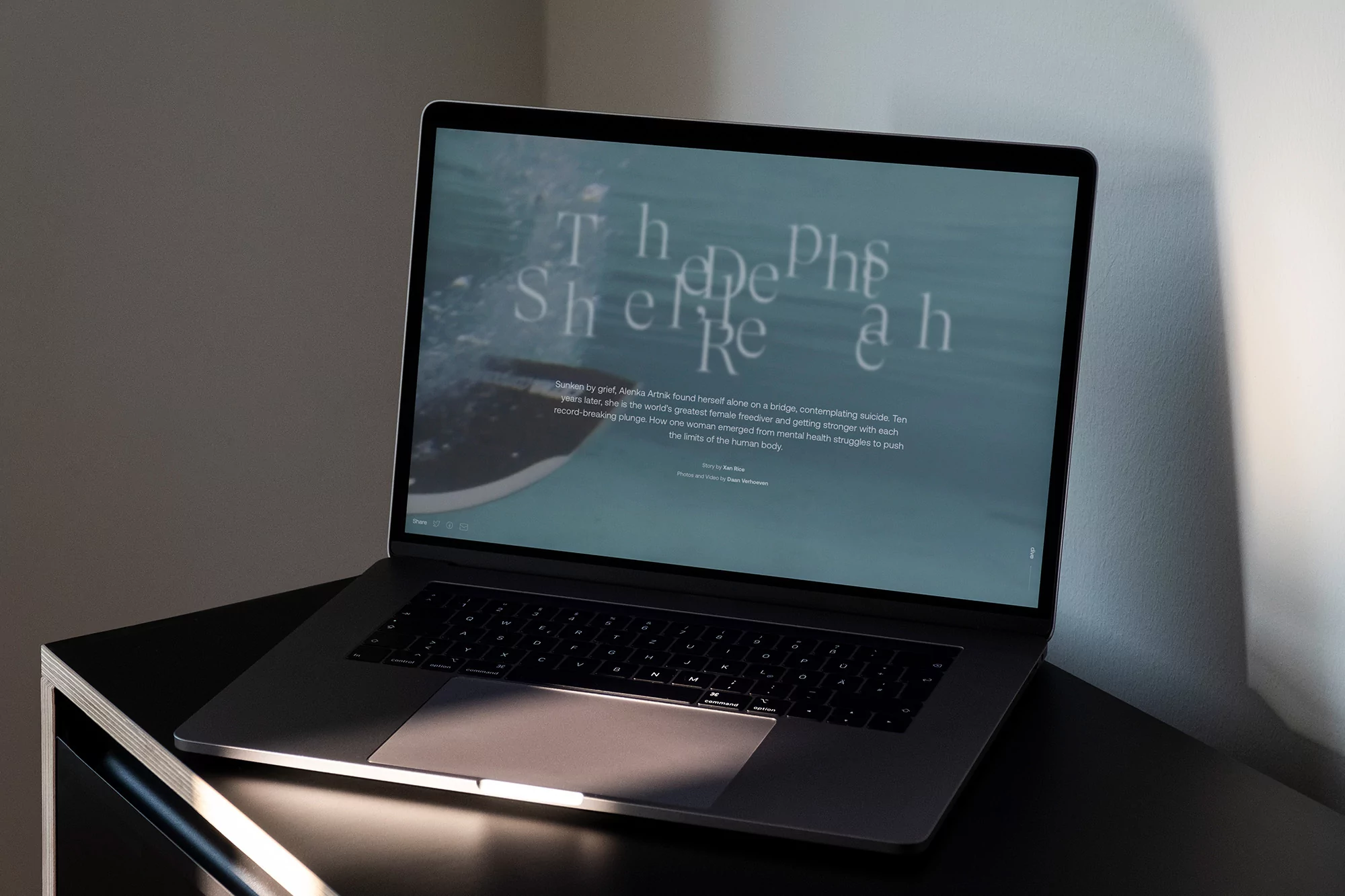 Laptop displays a website with teal water background and large text, resting on a sleek black surface near a light grey wall.