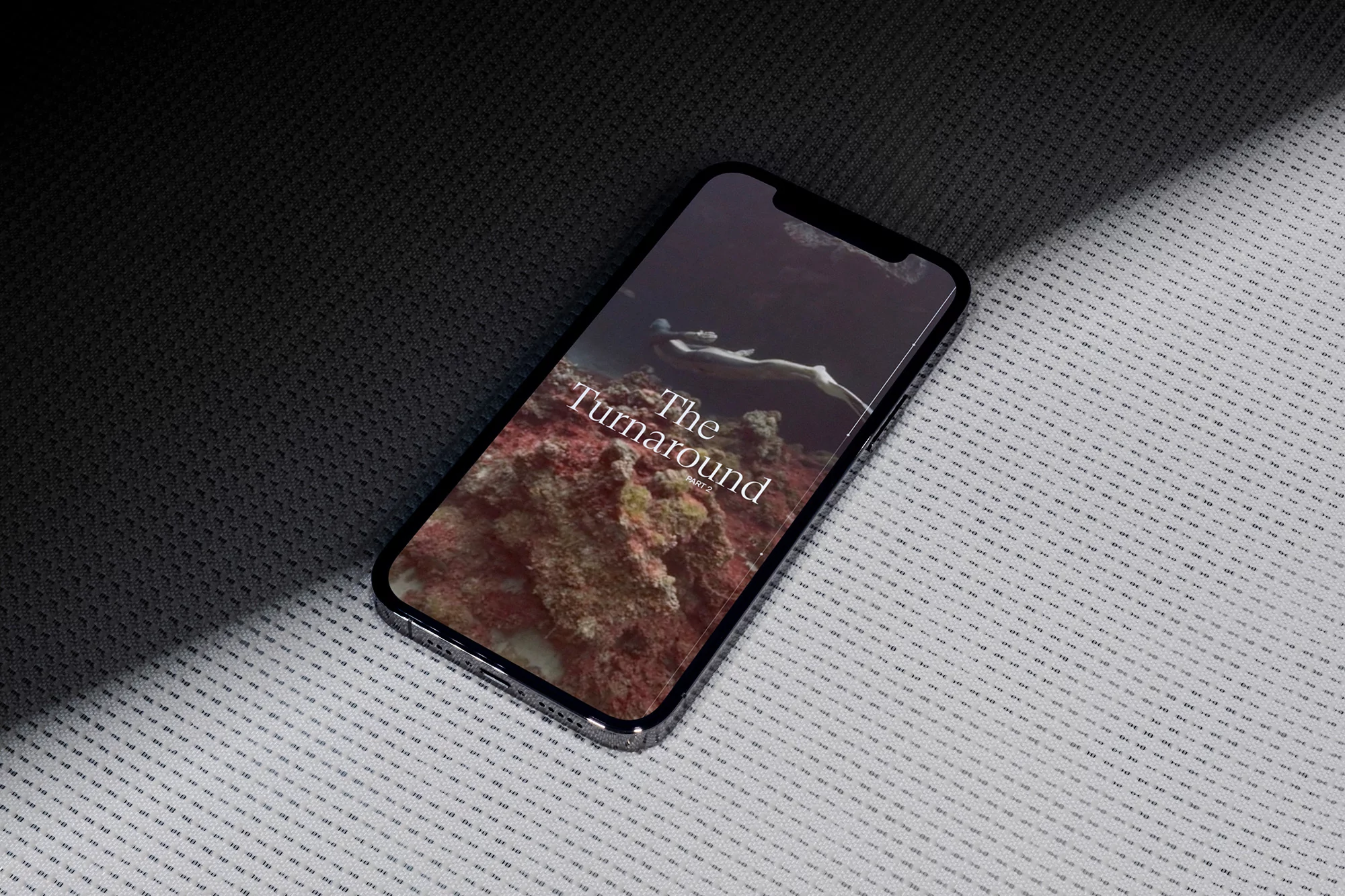 An iPhone displays a video of an underwater scene featuring a diver and coral, resting on a textured white and black surface with studio lighting.