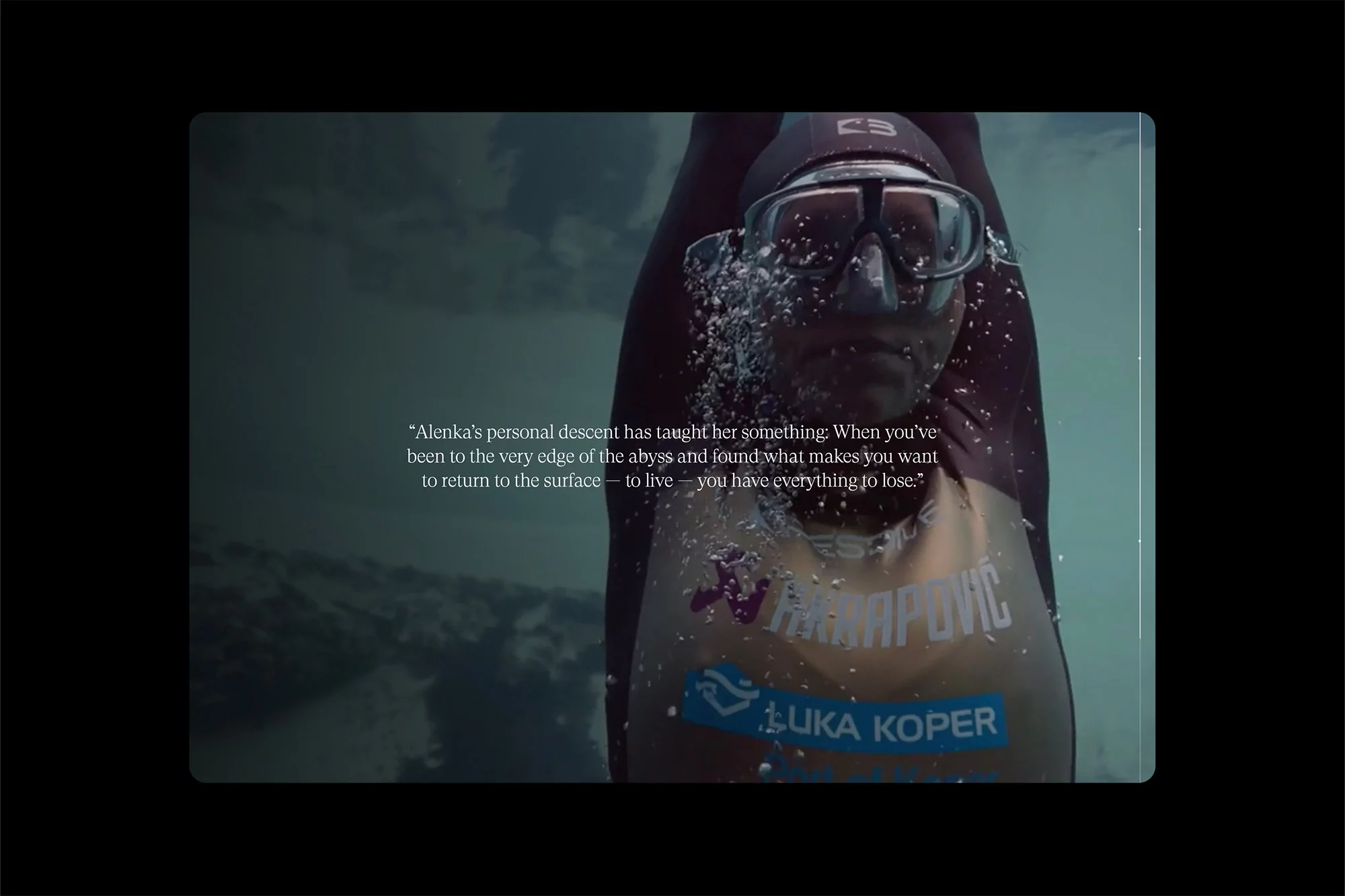 Underwater shot of a woman freediver wearing a wetsuit, goggles, and a cap, surrounded by bubbles in the water, focus on her face.