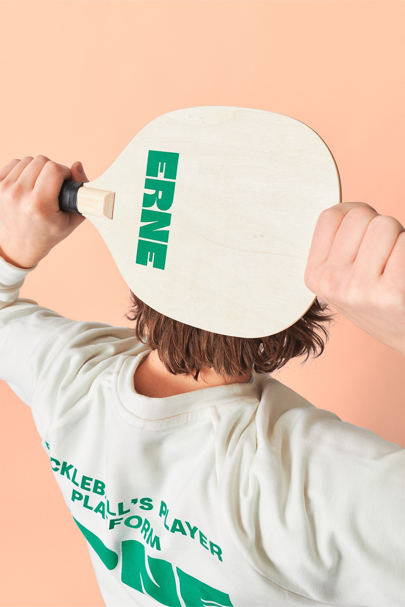 Person holds a wooden pickleball paddle with green text in front of their head indoors against a peach-colored background.