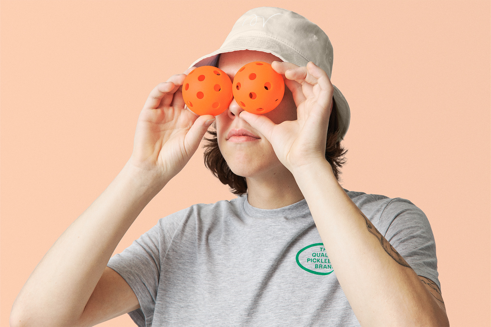 Person in bucket hat holds orange pickleballs over eyes in a studio shot against a peach background, wearing a gray graphic t-shirt.