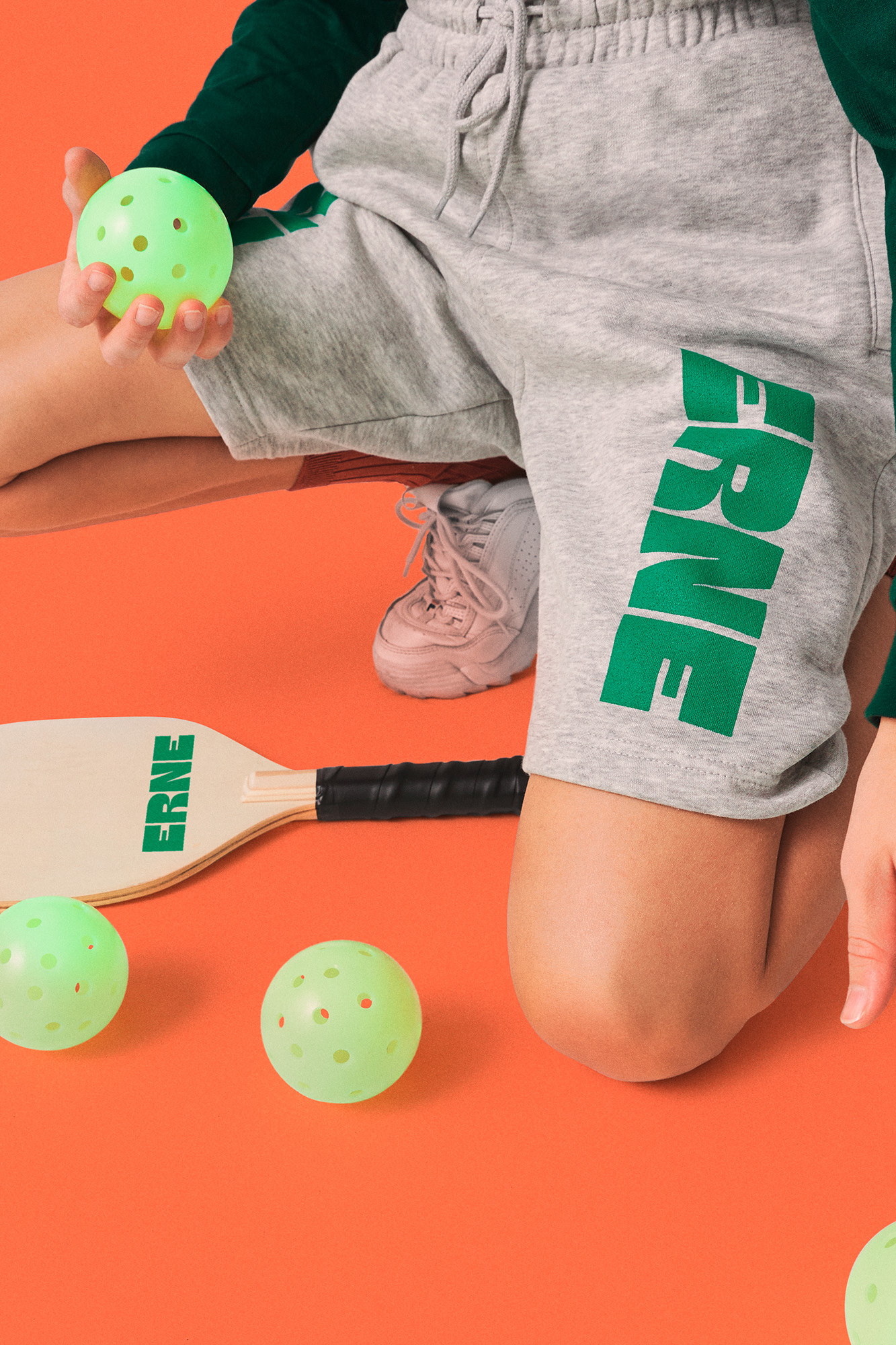 Child model displays gray ERNE-branded shorts, holding a perforated green pickleball, with a paddle on an orange backdrop.