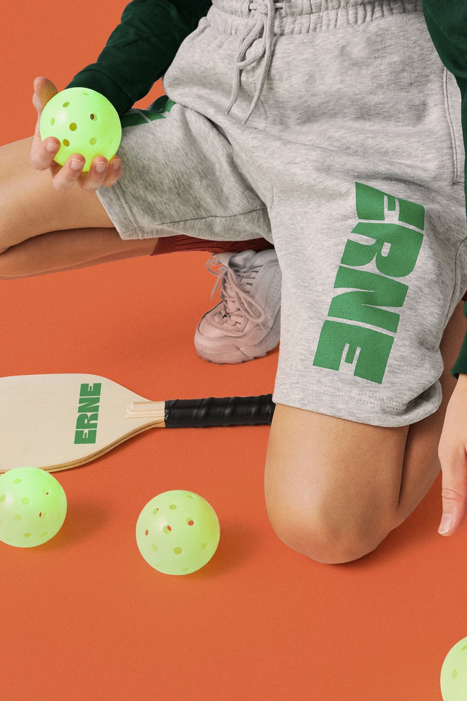 Close-up of a person in grey sweatshorts holding a bright green perforated pickleball, with a paddle and more balls on an orange floor