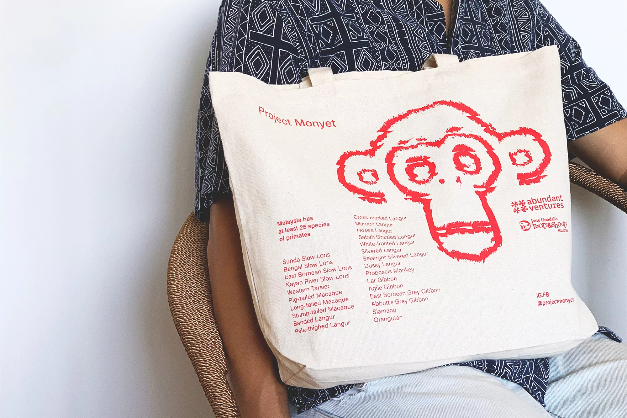 Close-up of a person holding a tote bag with a red monkey graphic and text. The bag lists primate species; person seated in wicker chair.