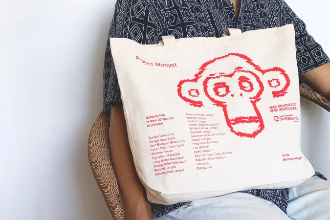 Close-up of a person holding a tote bag with a red monkey graphic and text. The bag lists primate species; person seated in wicker chair.