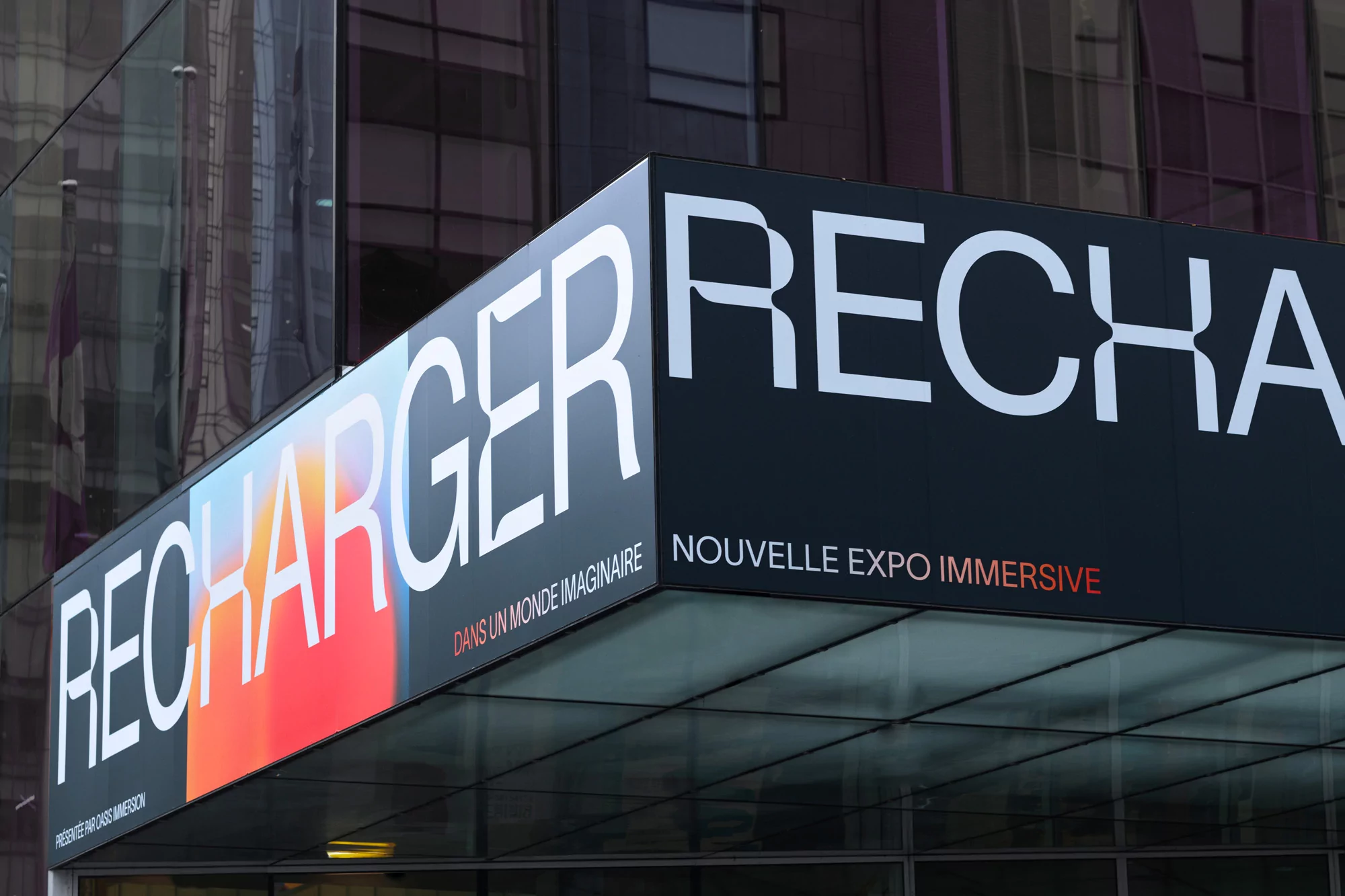 Architectural view of a modern building entrance with a large promotional sign for the 'RECHARGER' immersive expo.