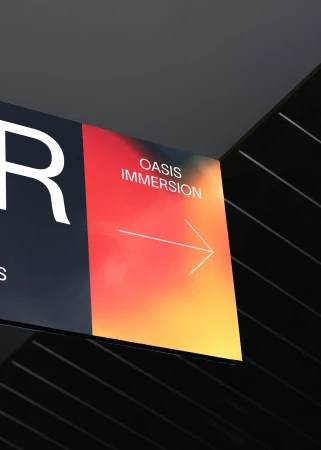 Directional sign with 'Oasis Immersion' in vibrant orange tones, featuring a white arrow against a dark, slatted ceiling for wayfinding.