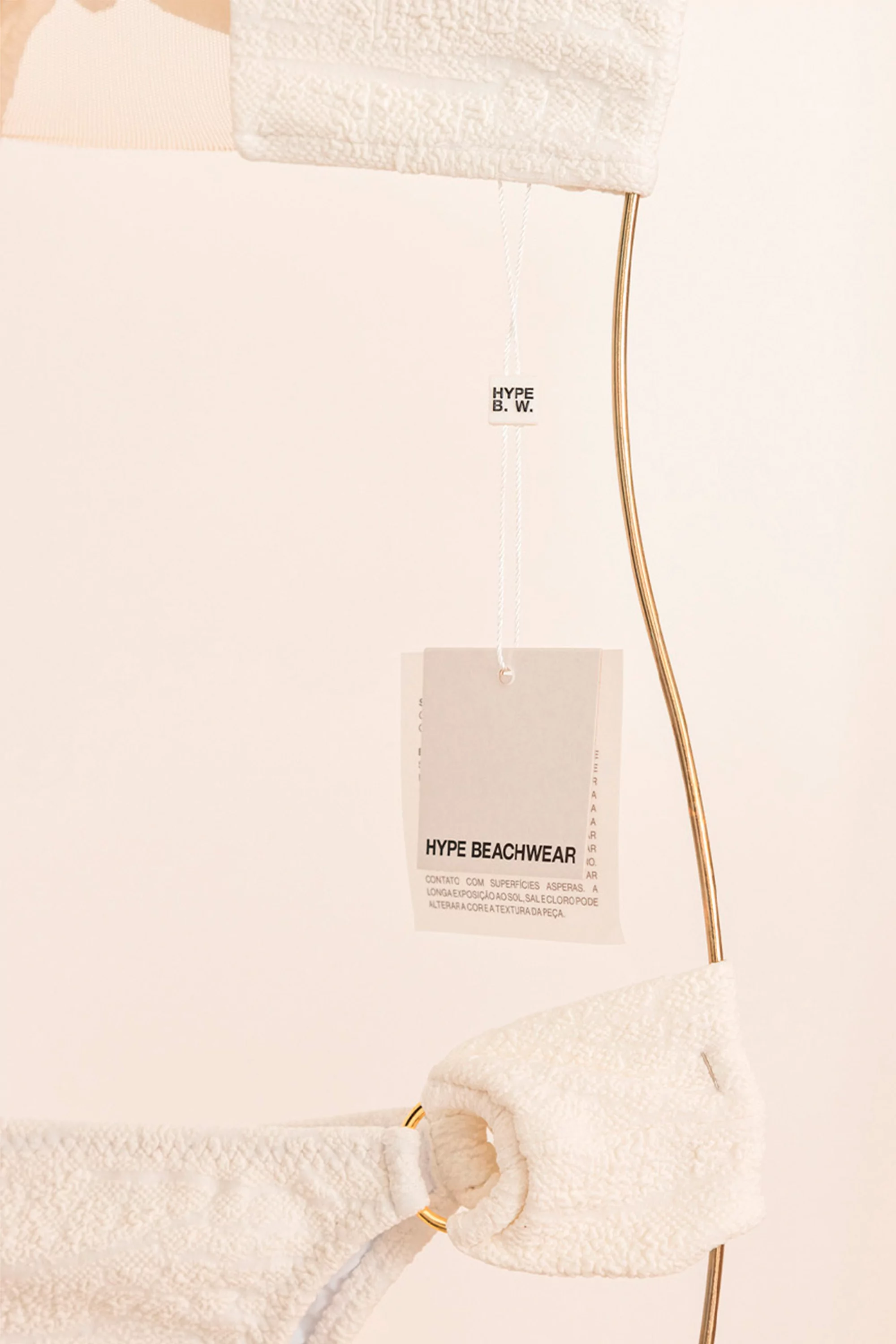 A textured white two-piece swimsuit with gold rings is displayed against a cream background with a garment tag for Hype Beachwear.