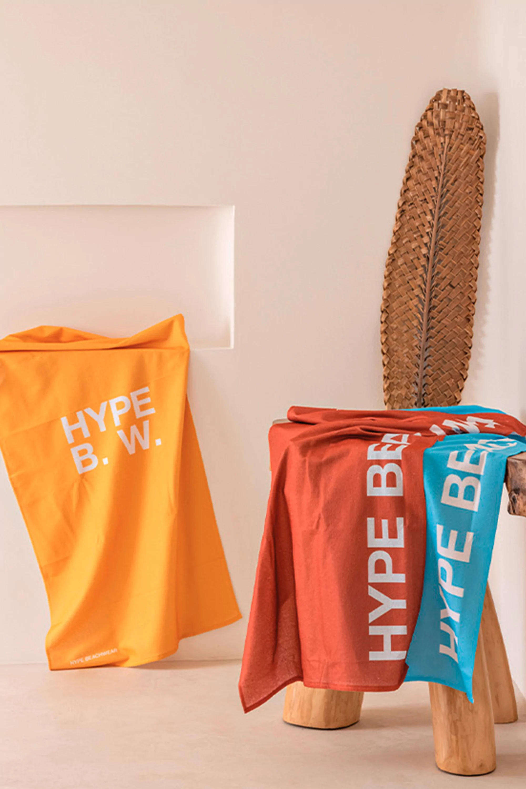 Three colorful towels with 'Hype B.W.' text displayed indoors on a wooden stool with a woven decor piece, minimalist background.
